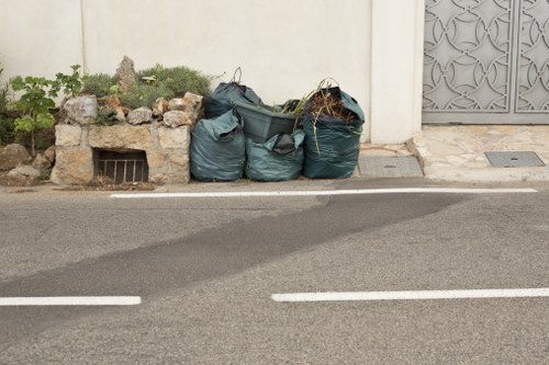 A collection of three dark green refuse bags filled with mixed waste, placed on a paved street surface near a white wall. The bags are crumpled and slightly open at the top, with some contents visible, such as plant debris and possibly household rubbish. Behind the bags, there is a small raised garden bed constructed of uneven stones, containing a few green plants and purple flowering bushes. To the right, a metal door with decorative cut-out patterns is set into the wall, which appears clean and well-maintained. The street has a smooth asphalt surface with white road markings visible in the foreground, indicating it is a public road likely in a residential or commercial area. The scene suggests a typical waste collection point where commercial waste removal services, such as those provided by Commercial Waste Removal Heston, may assist with rubbish clearance in the local vicinity near Heston, Middlesex.