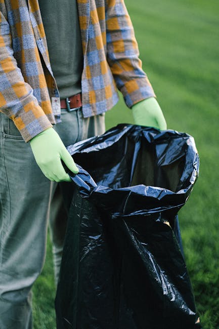 Two large black plastic garbage bags filled with waste are situated on the pavement next to a black metal fence, with a background of dense foliage and a building partially visible. The bags appear ti